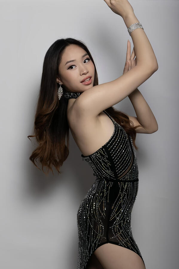 Angeli Chaves posing with arms raised in a black rhinestone halter gown against a gray backdrop