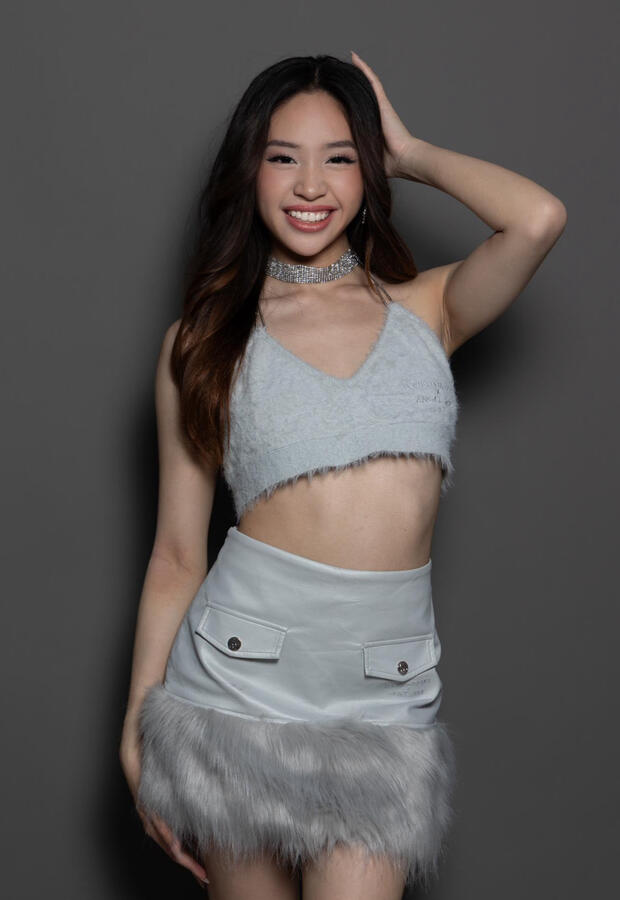 Angeli Chaves smiling confidently in a fuzzy gray crop top and leather mini skirt with fur trim, hand resting on her head