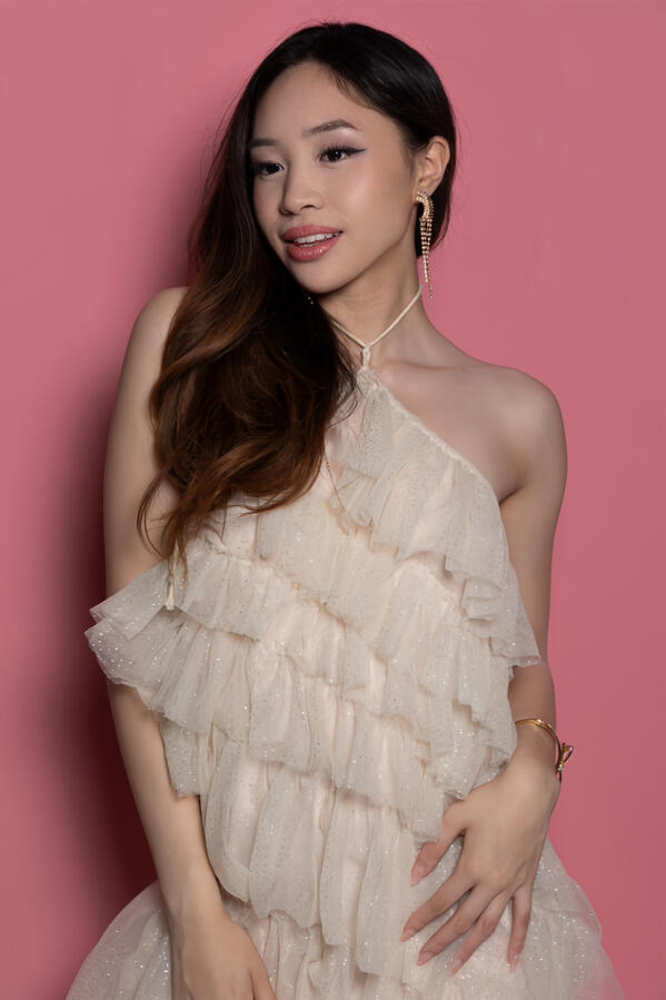 Angeli Chaves in a soft pink halter tiered ruffle dress with gold jewelry against a pink backdrop