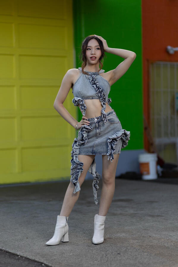 Angeli Chaves outdoors in a ruffled denim crop top and skirt with white heeled boots, posing against a colorful wall